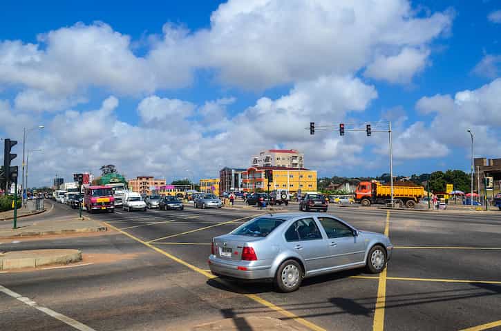 Uma estrada em Gana, repleta de carros sob um céu azul levemente nublado. Ao fundo, prédios e outros veículos estacionados.