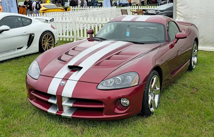 Um Dodge Viper vinho com faixas esportivas brancas parado sobre a grama.